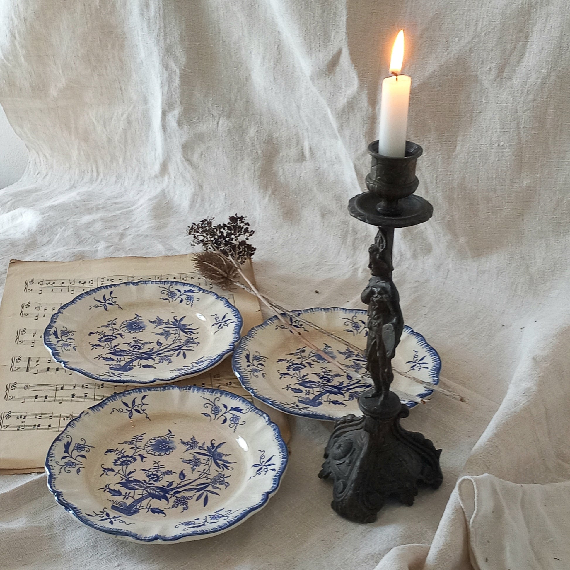 Three blue and white ceramic plates with a candlestick holder and lit candle on a textured white fabric background.