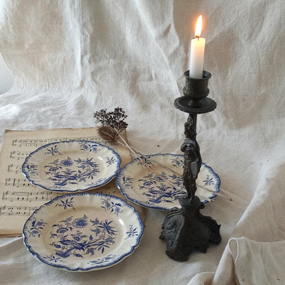 Three blue and white ceramic plates with a candlestick holder and lit candle on a textured white fabric background.