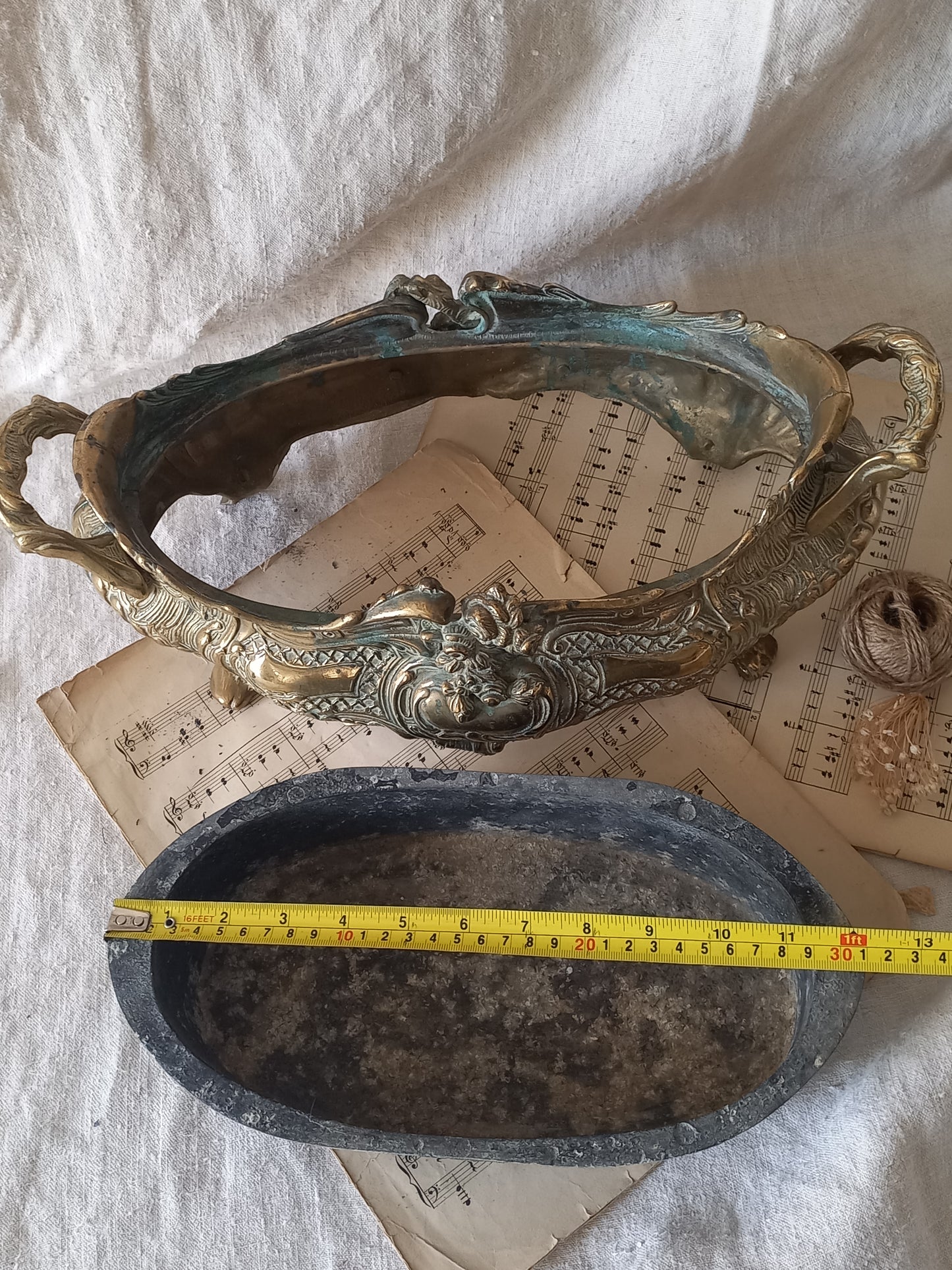 An ornate antique decorative planter resting on a vintage sheet music page against a draped white fabric background with a tape measure for reference.