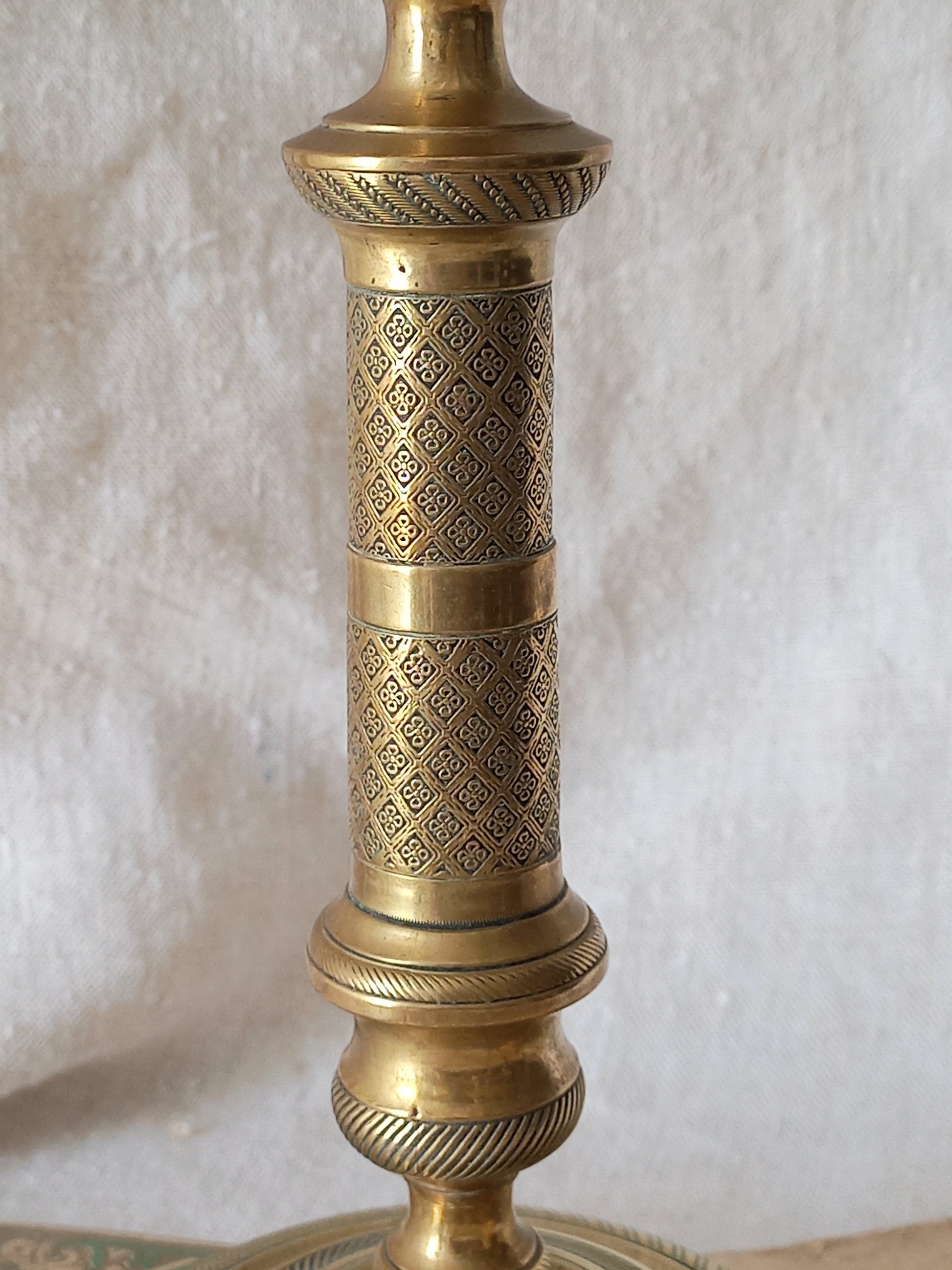 An ornate brass candlestick with intricate geometric cutout patterns on its cylindrical body, set against a neutral textured fabric background.