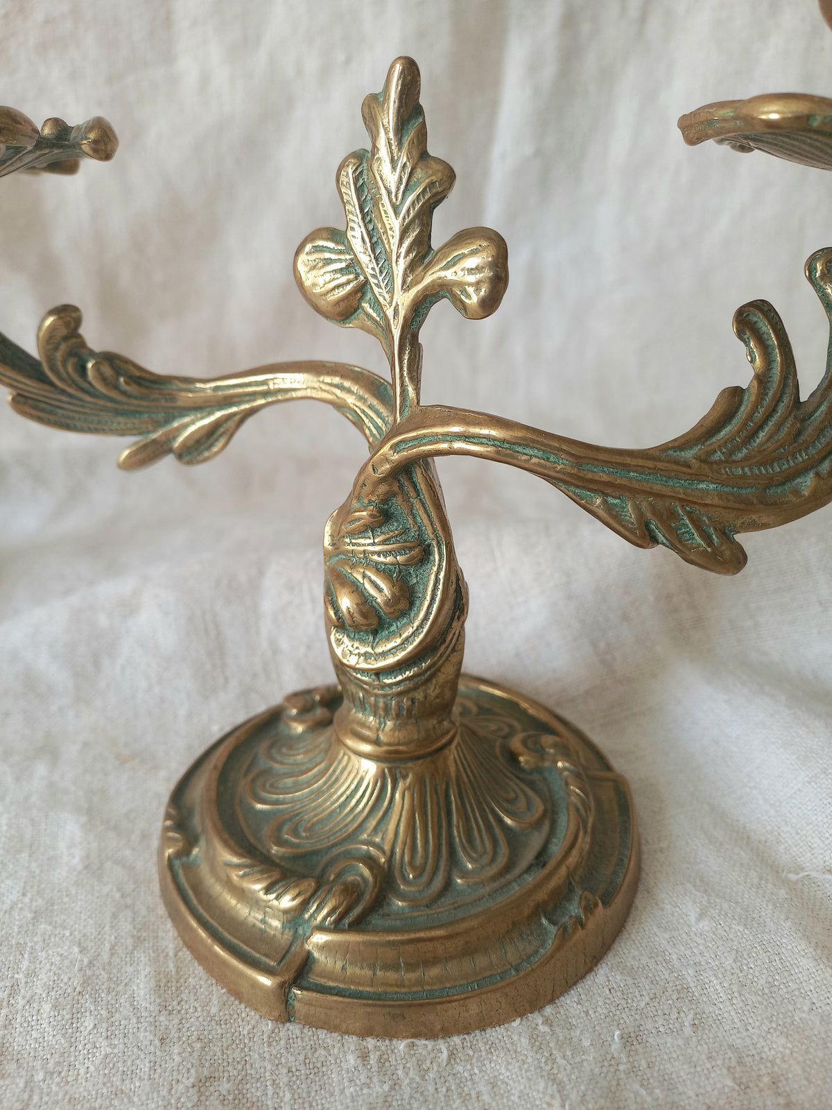 An ornate brass candelabra with a circular pedestal base and leaf-shaped arms, photographed against a draped white fabric background.