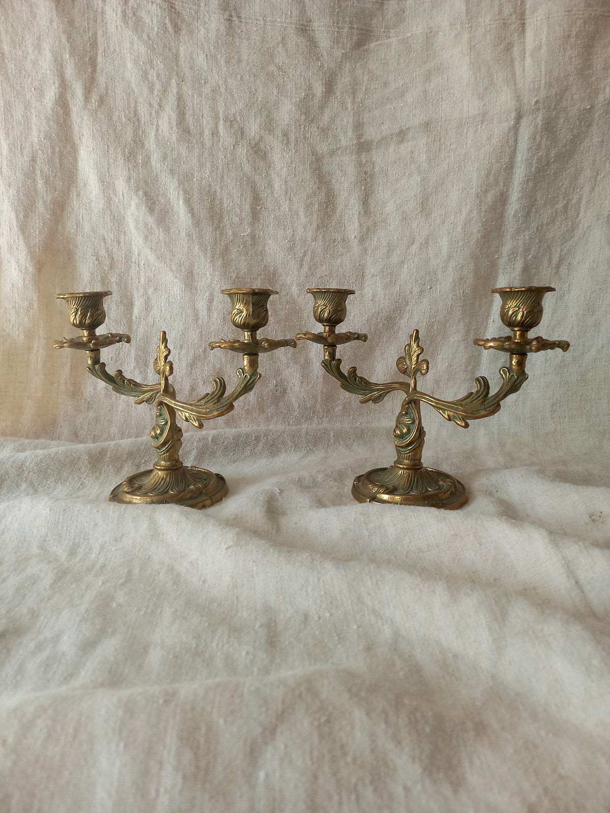 A pair of ornate brass candelabras displayed on a draped white fabric background.