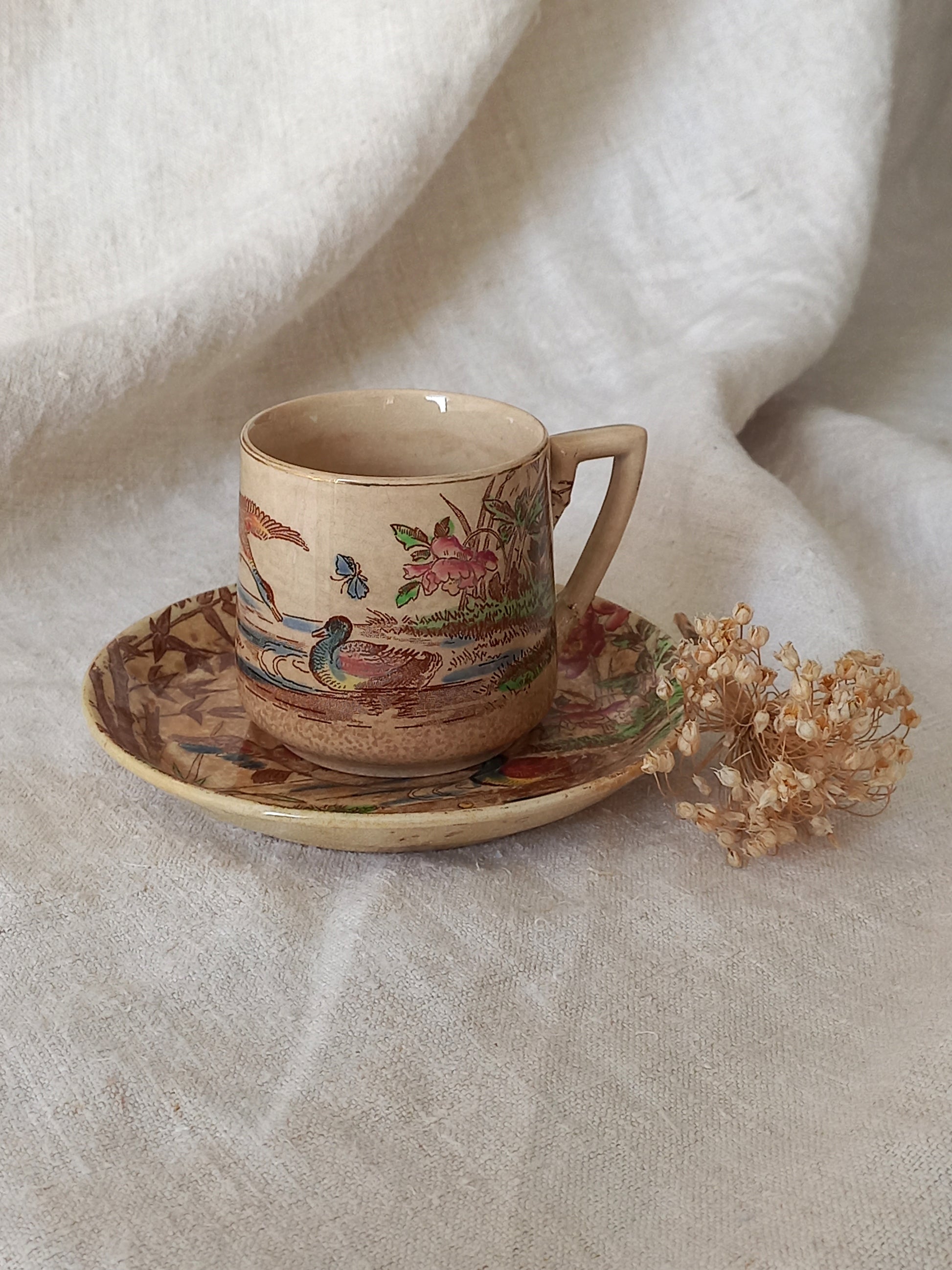 Vintage ceramic cup and saucer with nature-themed design on a textured fabric background