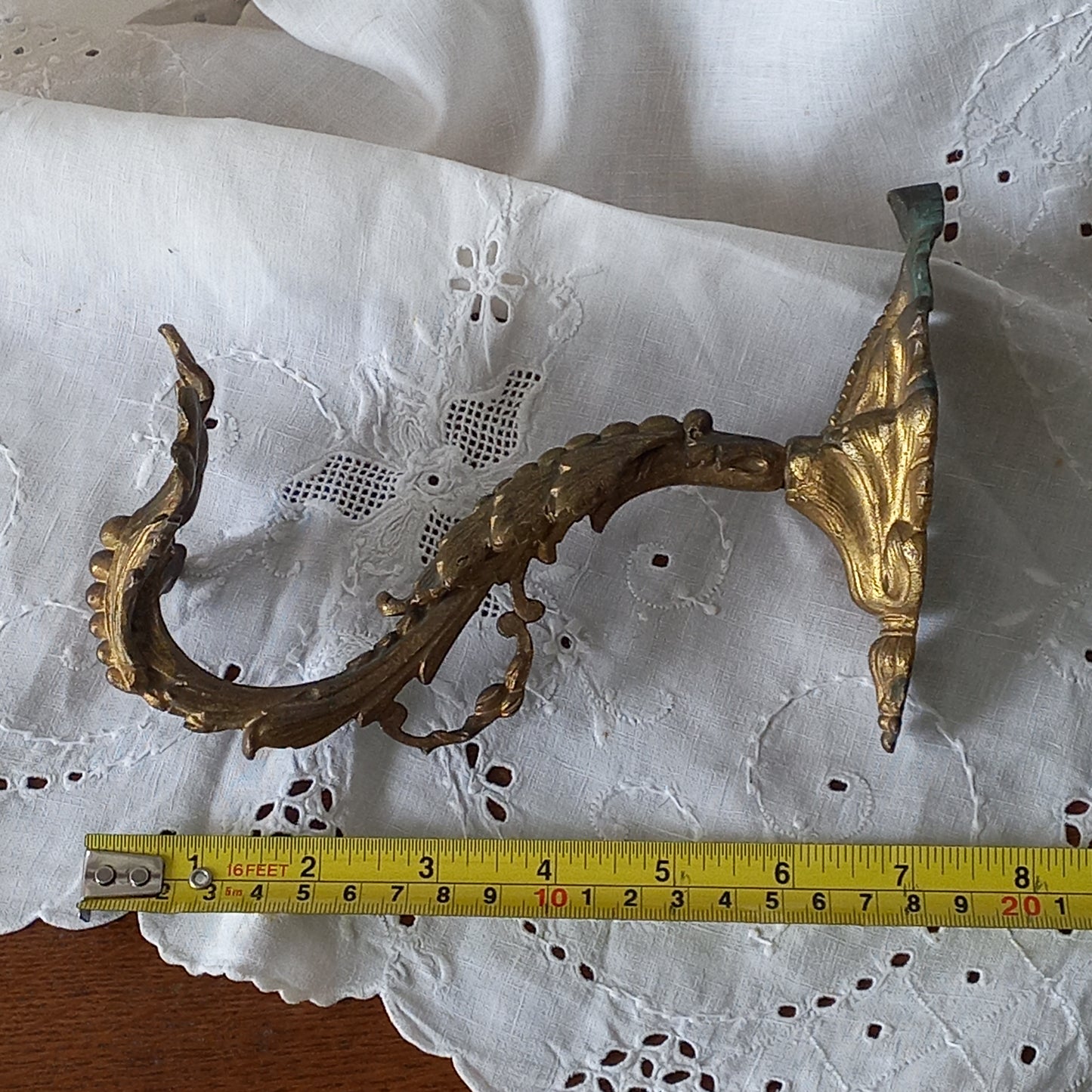 Decorative metal wall bracket with gold accents on a white lace background, measured with a tape measure.