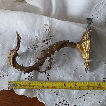Decorative metal wall bracket with gold accents on a white lace background, measured with a tape measure.