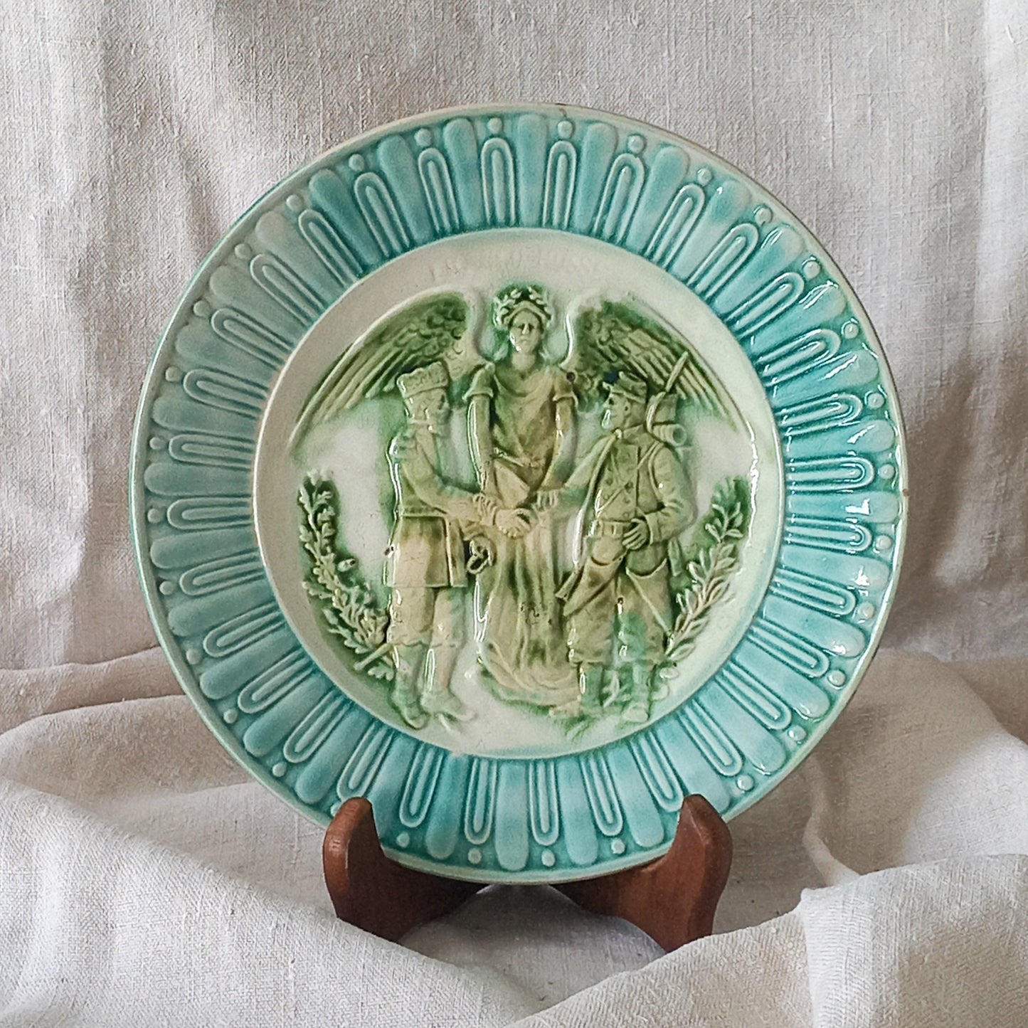 A French antique decorative majolica plate with stunning blue/green colours and an interesting historical background.

This lovely old plate was manufactured as a commemorative piece to celebrate the Franco-Russian Alliance made by the French Republic and the Russian Empire between 1891-1894 (military co-operation agreements) – and that lasted until 1917. The plate depicts a Russian and French soldier shaking hands with a winged female angel in between them.