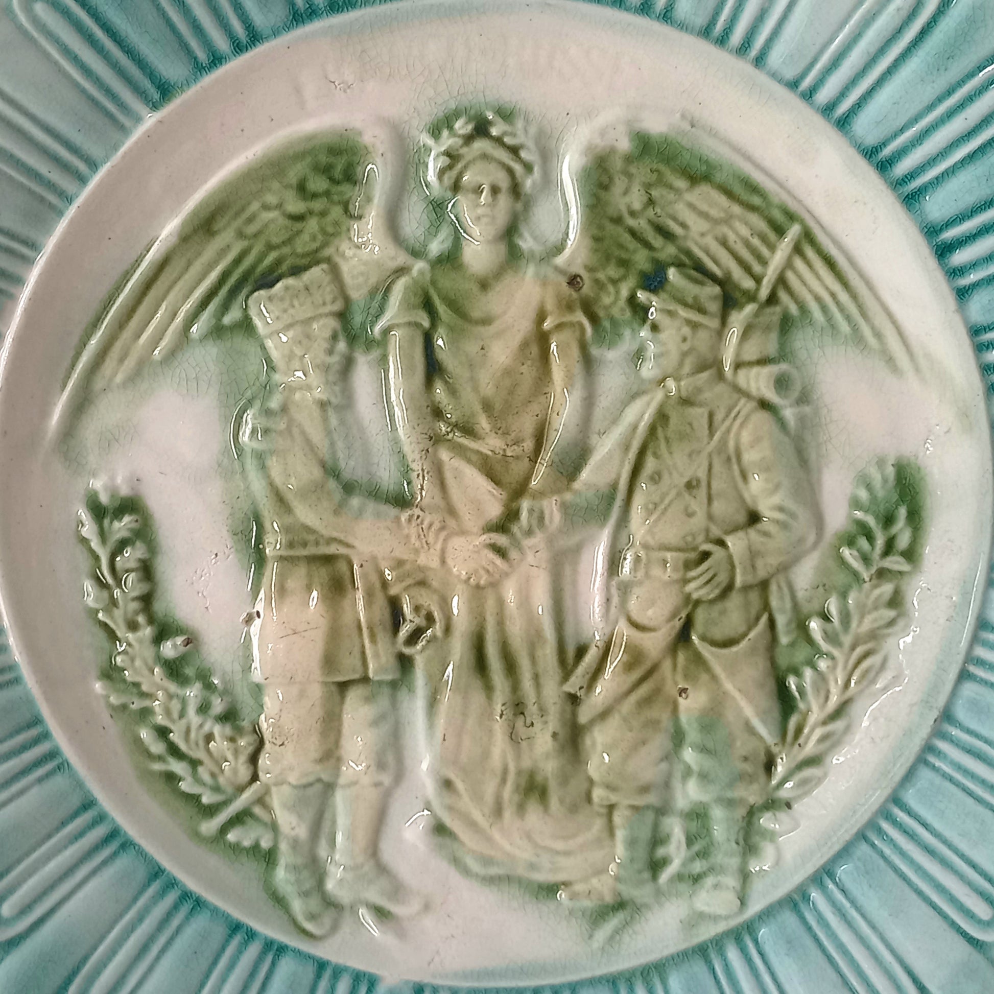 A French antique decorative majolica plate with stunning blue/green colours and an interesting historical background.

This lovely old plate was manufactured as a commemorative piece to celebrate the Franco-Russian Alliance made by the French Republic and the Russian Empire between 1891-1894 (military co-operation agreements) – and that lasted until 1917. The plate depicts a Russian and French soldier shaking hands with a winged female angel in between them.