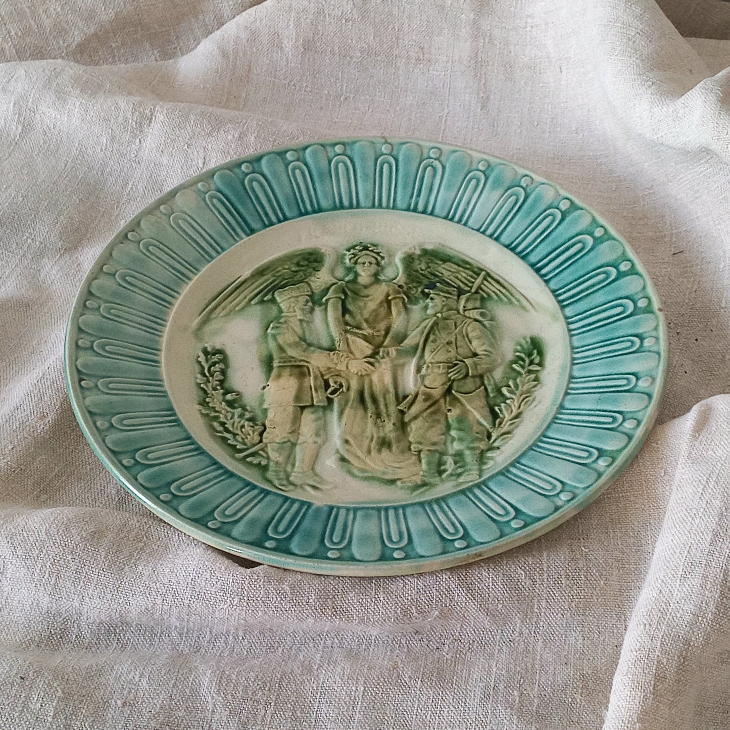 A French antique decorative majolica plate with stunning blue/green colours and an interesting historical background.

This lovely old plate was manufactured as a commemorative piece to celebrate the Franco-Russian Alliance made by the French Republic and the Russian Empire between 1891-1894 (military co-operation agreements) – and that lasted until 1917. The plate depicts a Russian and French soldier shaking hands with a winged female angel in between them.