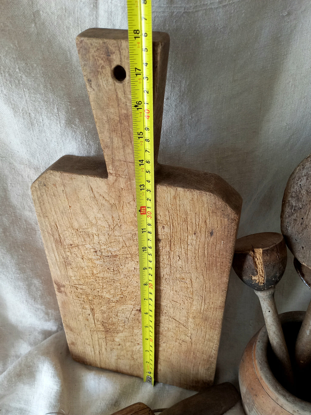 A rustic wooden cutting board with a handle and a hole near the top, shown standing upright with a yellow tape measure placed vertically along its center. The board is set against a draped beige fabric backdrop, with other vintage kitchen items partially visible on the right.