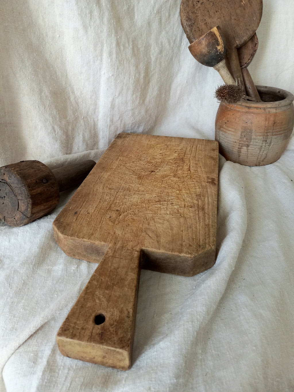A rustic wooden cutting board with a handle, displayed on draped white linen fabric alongside other vintage wooden kitchen utensils.