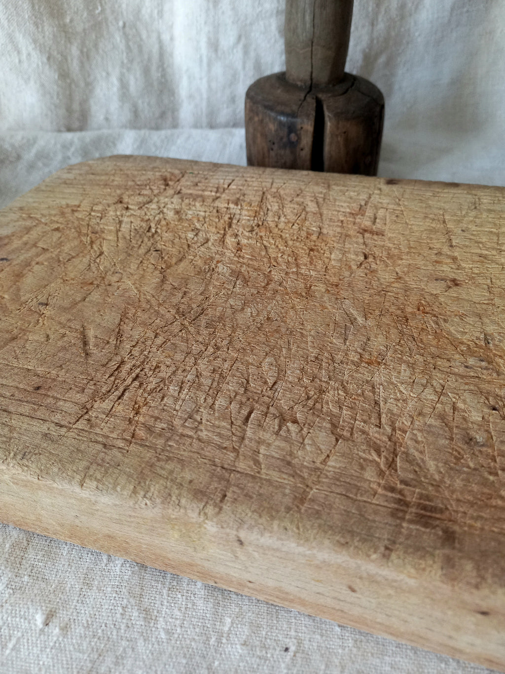 A close-up of a heavily used, rustic wooden cutting board with deep knife scratches, resting on a white linen cloth with a dark wooden mallet in the background.