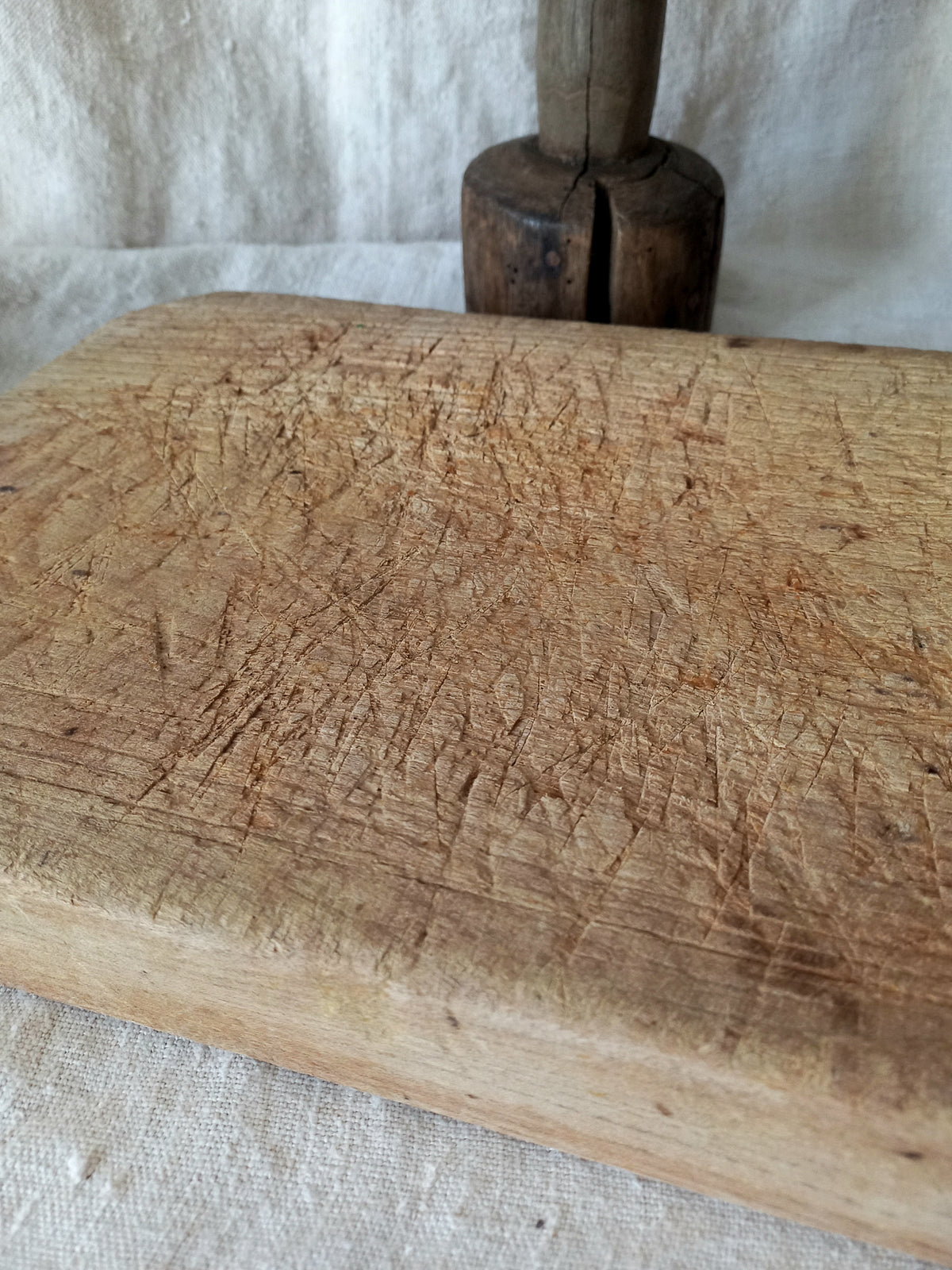 A close-up of a heavily used, rustic wooden cutting board with deep knife scratches, resting on a white linen cloth with a dark wooden mallet in the background.