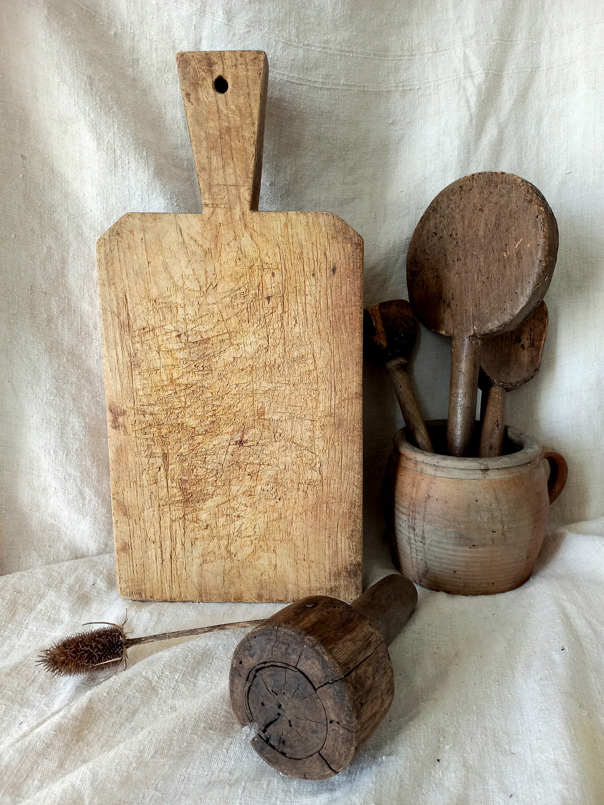 A rustic wooden cutting board with a handle stands upright next to a small clay pot holding wooden spoons, with a wooden pestle and a small brush resting on a draped white fabric background.
