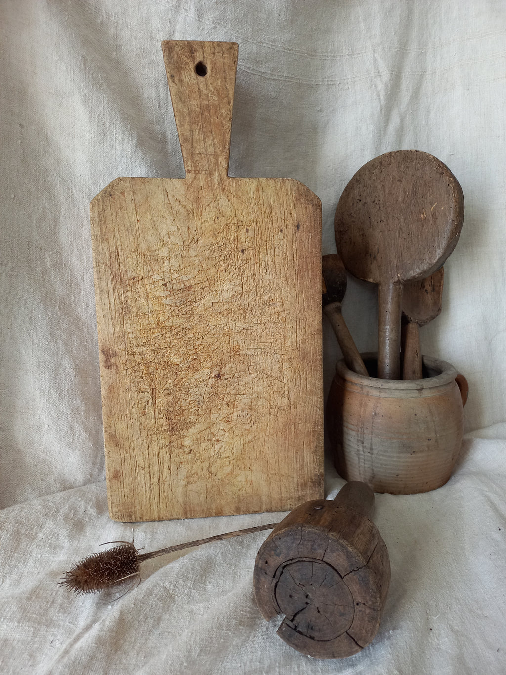 A vintage wooden cutting board with a handle, shown alongside a wooden rolling pin and a small cleaning brush on a white fabric background.