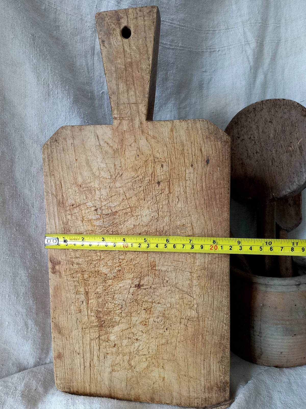 A rustic wooden cutting board with a handle and a hole near the top, shown with a yellow tape measure across the middle and a ceramic container in the background.