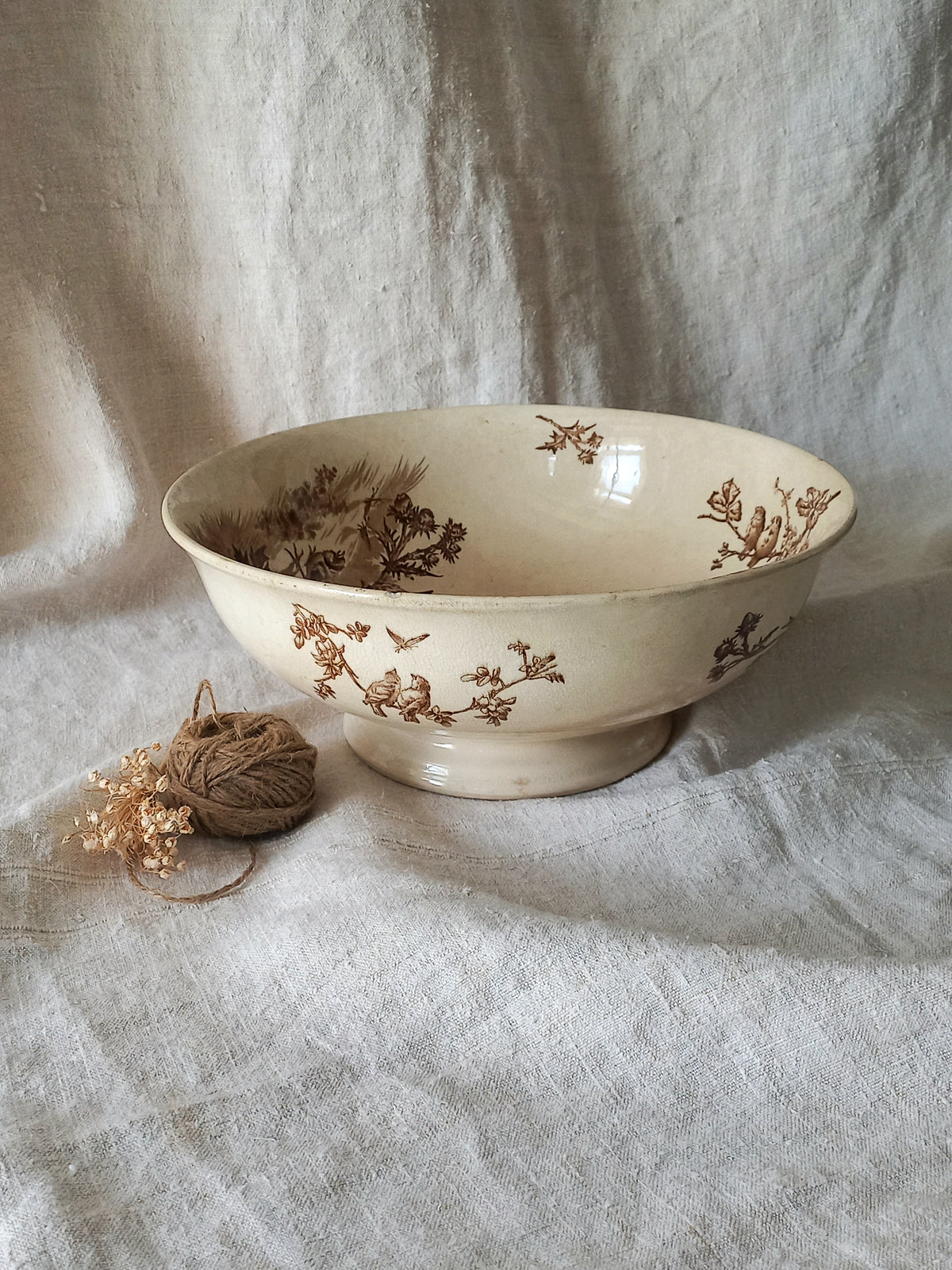 An off-white ceramic bowl with a brown floral and bird pattern, styled on draped linen fabric next to a small ball of twine and dried flowers.