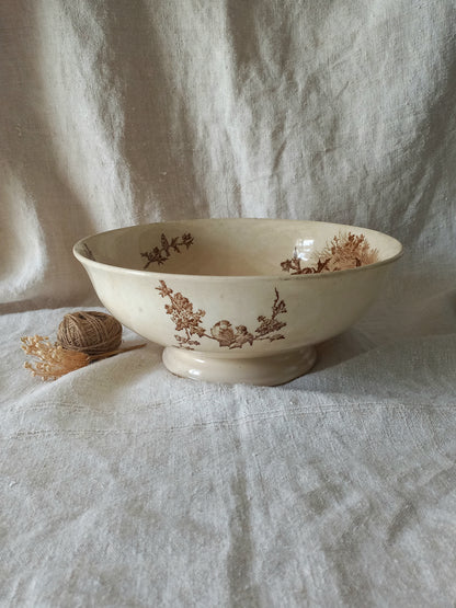 An off-white ceramic bowl with a brown floral toile pattern, set on a draped linen fabric background next to a small decorative ball of twine.