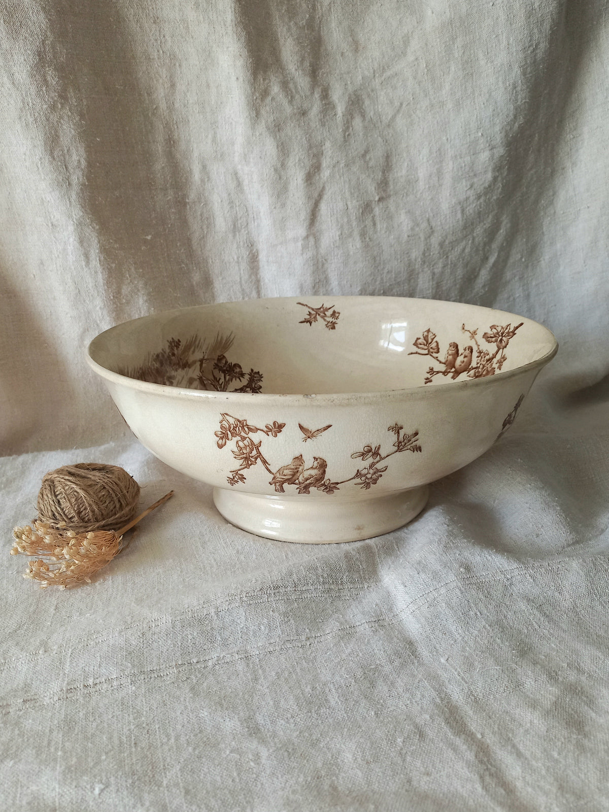 An off-white ceramic bowl with a brown floral and bird pattern, set against a draped linen fabric backdrop with a small ball of twine prop on the left.