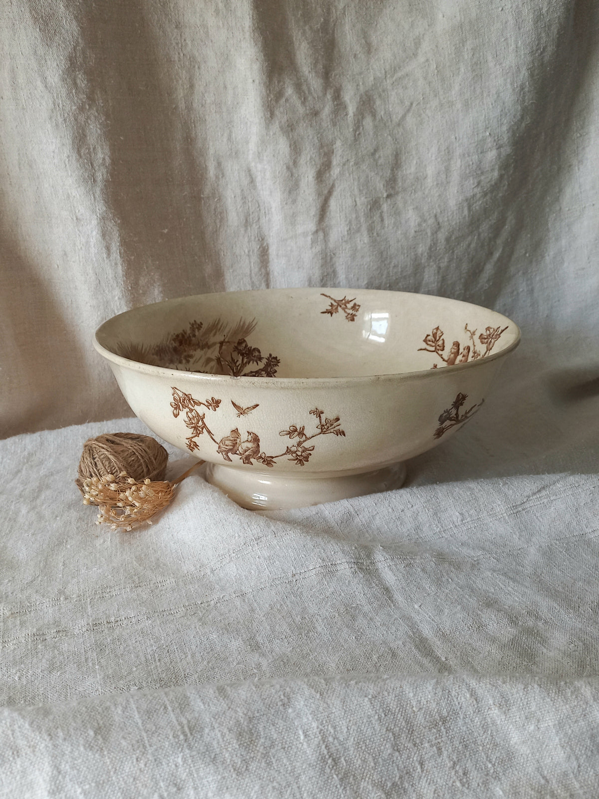 An off-white ceramic bowl with a brown floral and bird pattern, styled on a textured white linen fabric backdrop next to a small ball of twine.