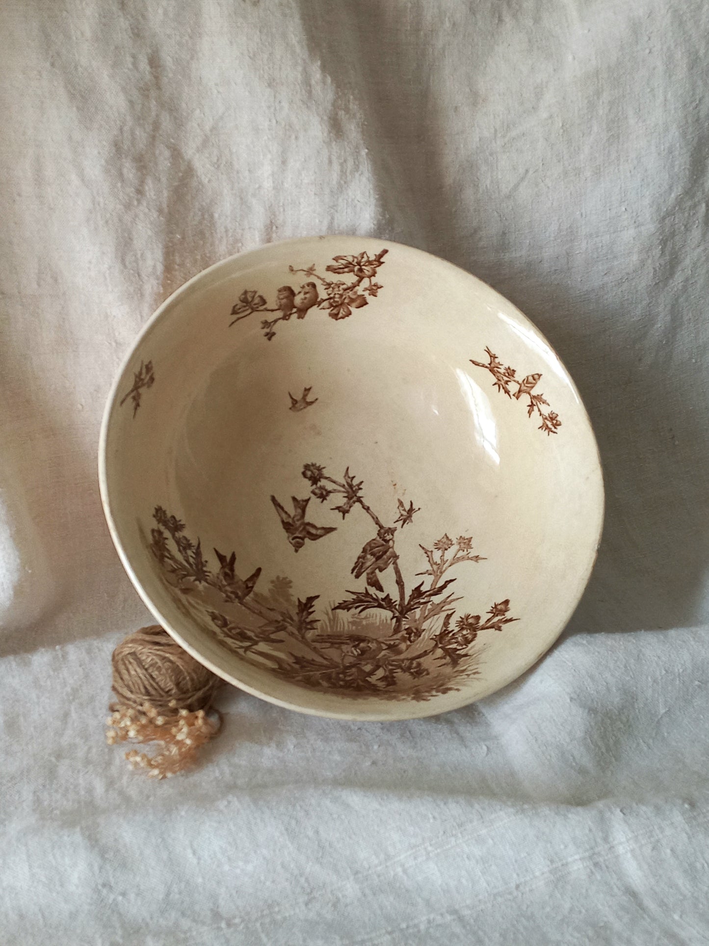 An off-white ceramic bowl with a brown sepia floral and bird pattern, resting on a textured white fabric background next to a small decorative object.