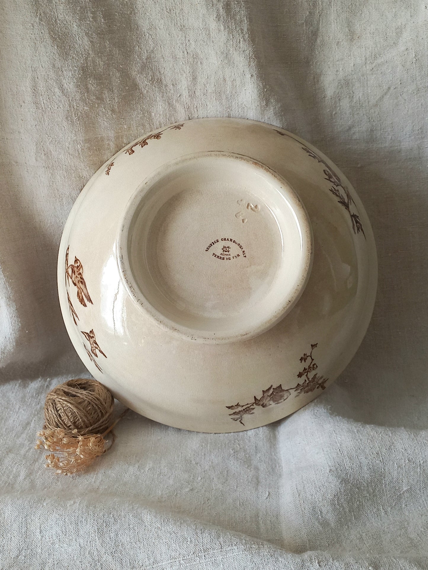 An antique cream-colored ceramic bowl with brown floral illustrations, shown upside down on a draped white linen fabric with a small ball of twine in the foreground.