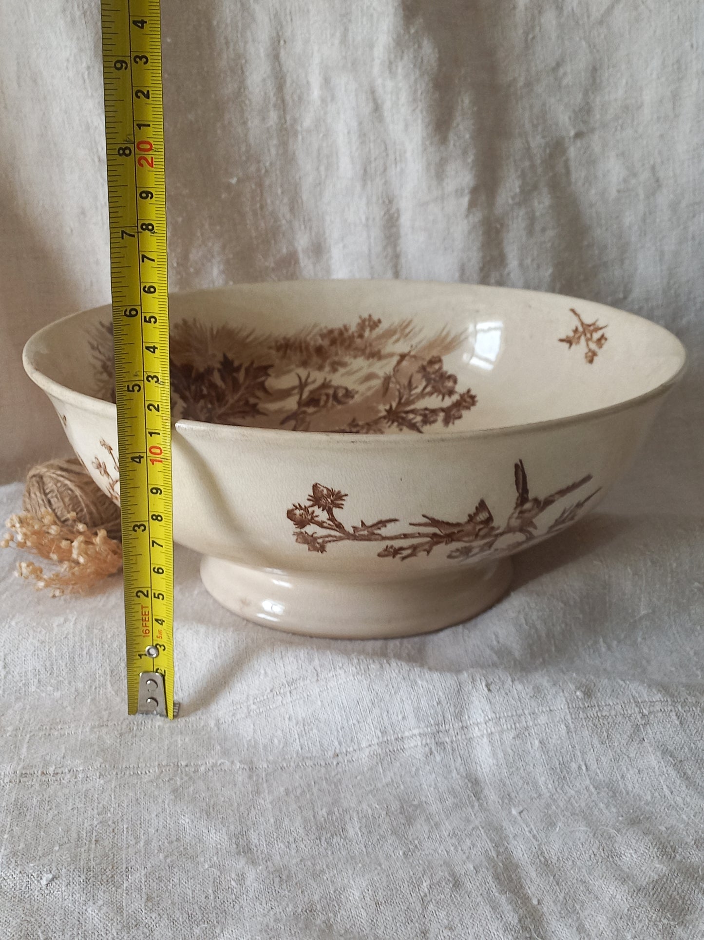 An off-white ceramic bowl with a brown sepia landscape and bird pattern, shown with a yellow tape measure standing next to it on a white fabric background.