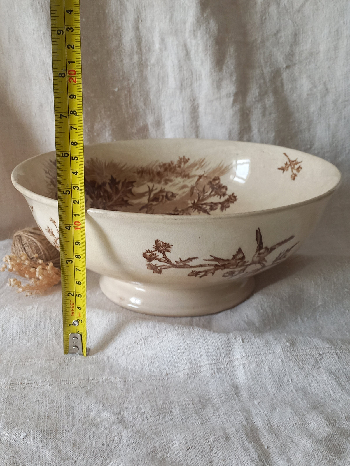 An off-white ceramic bowl with a brown sepia landscape and bird pattern, shown with a yellow tape measure standing next to it on a white fabric background.