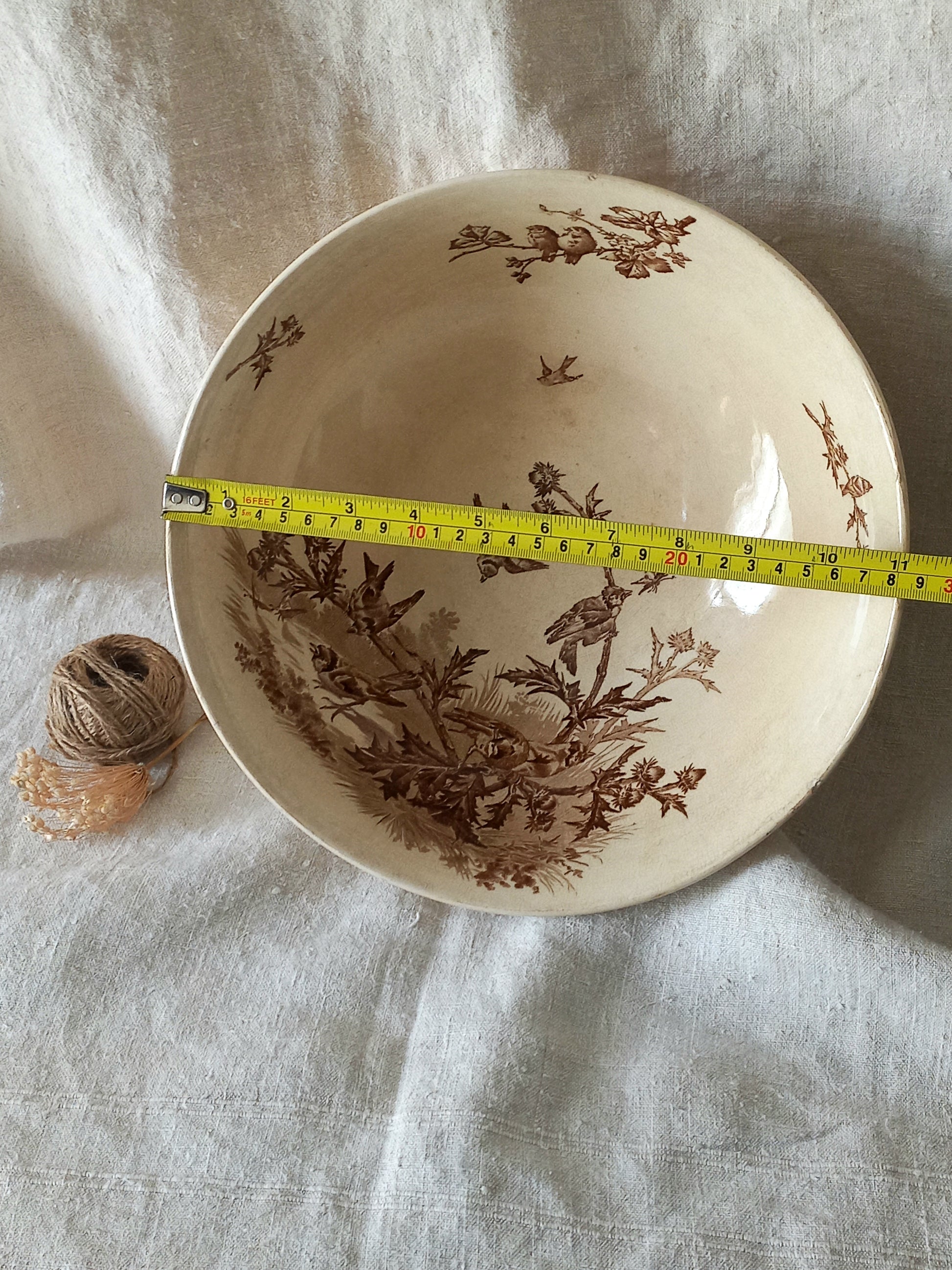 An off-white ceramic bowl with a brown sepia floral and bird pattern, shown with a yellow tape measure across the rim to indicate size, set on a textured white fabric background.