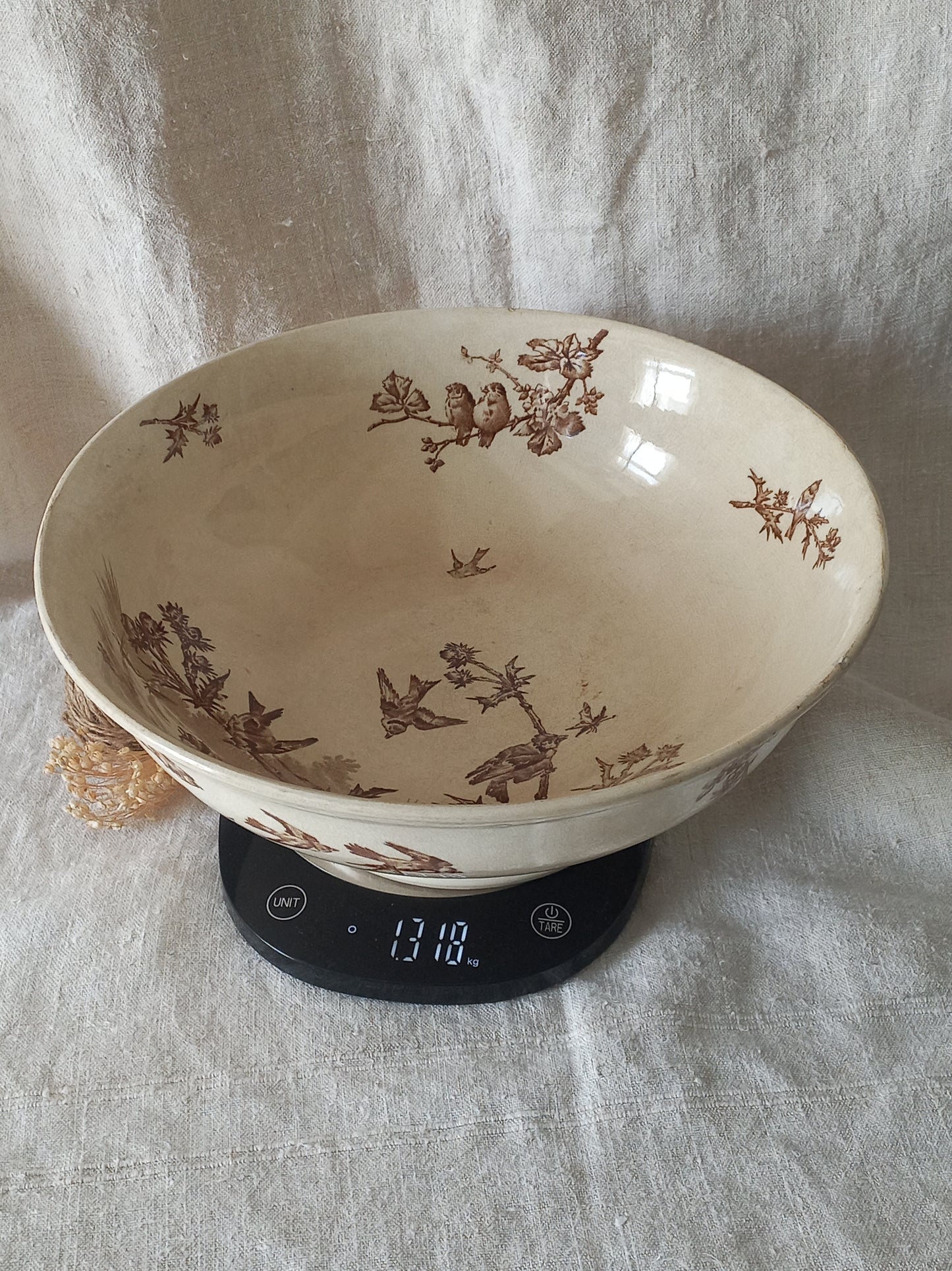 A large cream-colored ceramic bowl with brown floral and bird motifs, shown resting on a digital kitchen scale against a draped fabric backdrop.