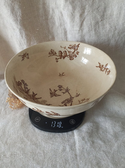 A large cream-colored ceramic bowl with brown floral and bird motifs, shown resting on a digital kitchen scale against a draped fabric backdrop.