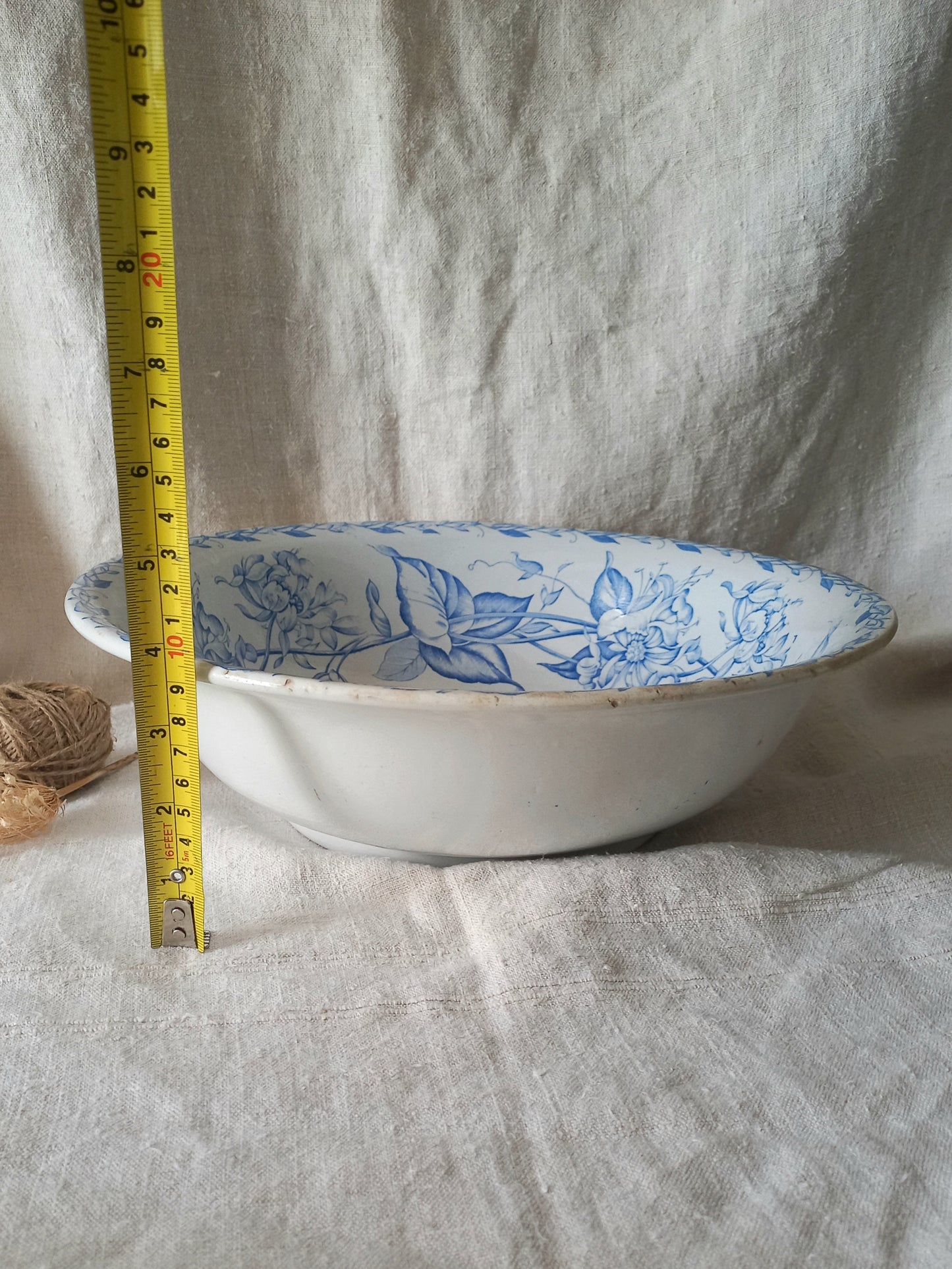 A white ceramic bowl with a blue floral pattern inside, shown next to a yellow tape measure for scale against a draped white linen background.