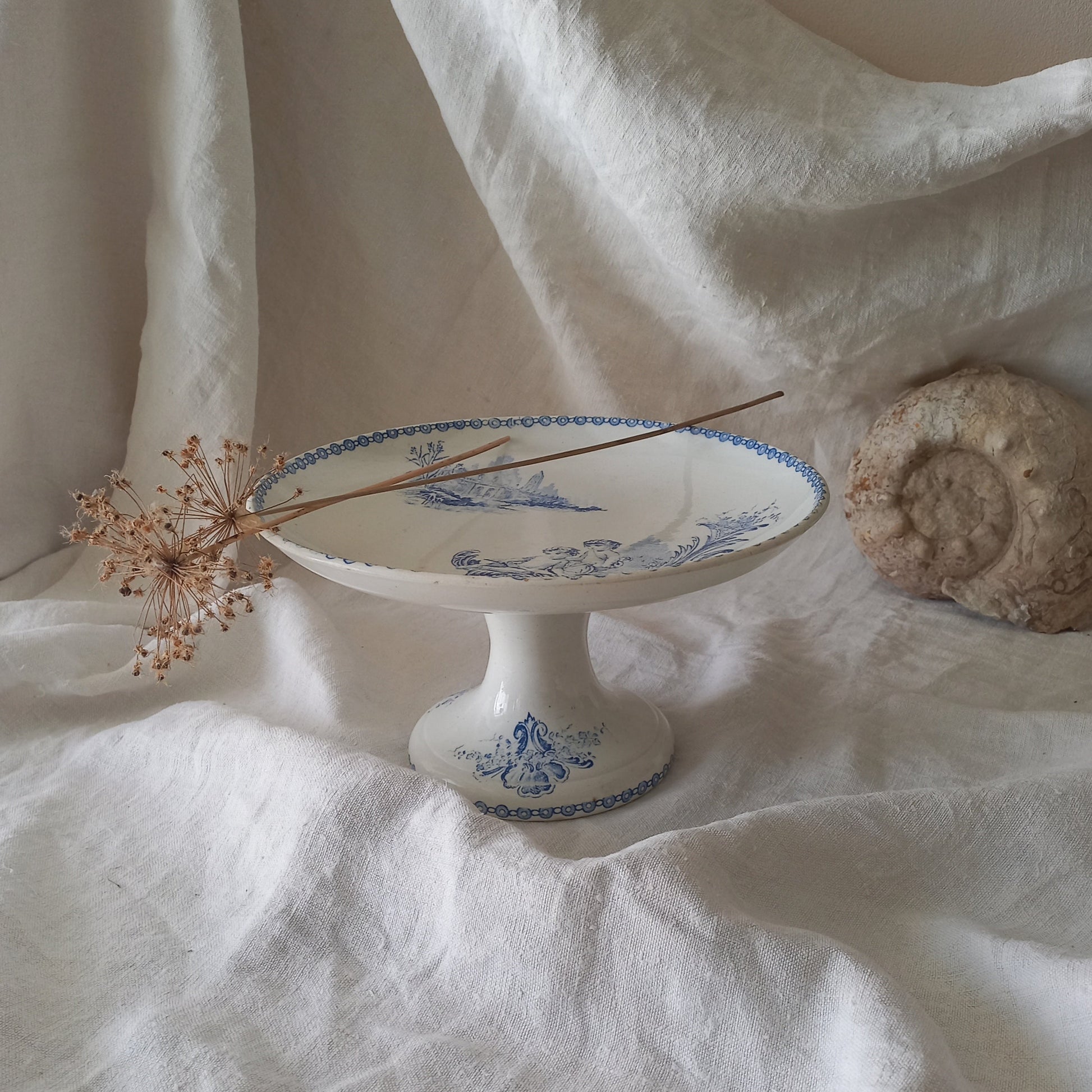 An attractive French antique footed ironstone cake plate with a blue transfer pattern depicting architecture, cherubs, and flowers; a very pretty and delicate pattern.

Original antique ironstone pieces of this age and condition are becoming increasingly difficult to source to this a rare treat to discover.

