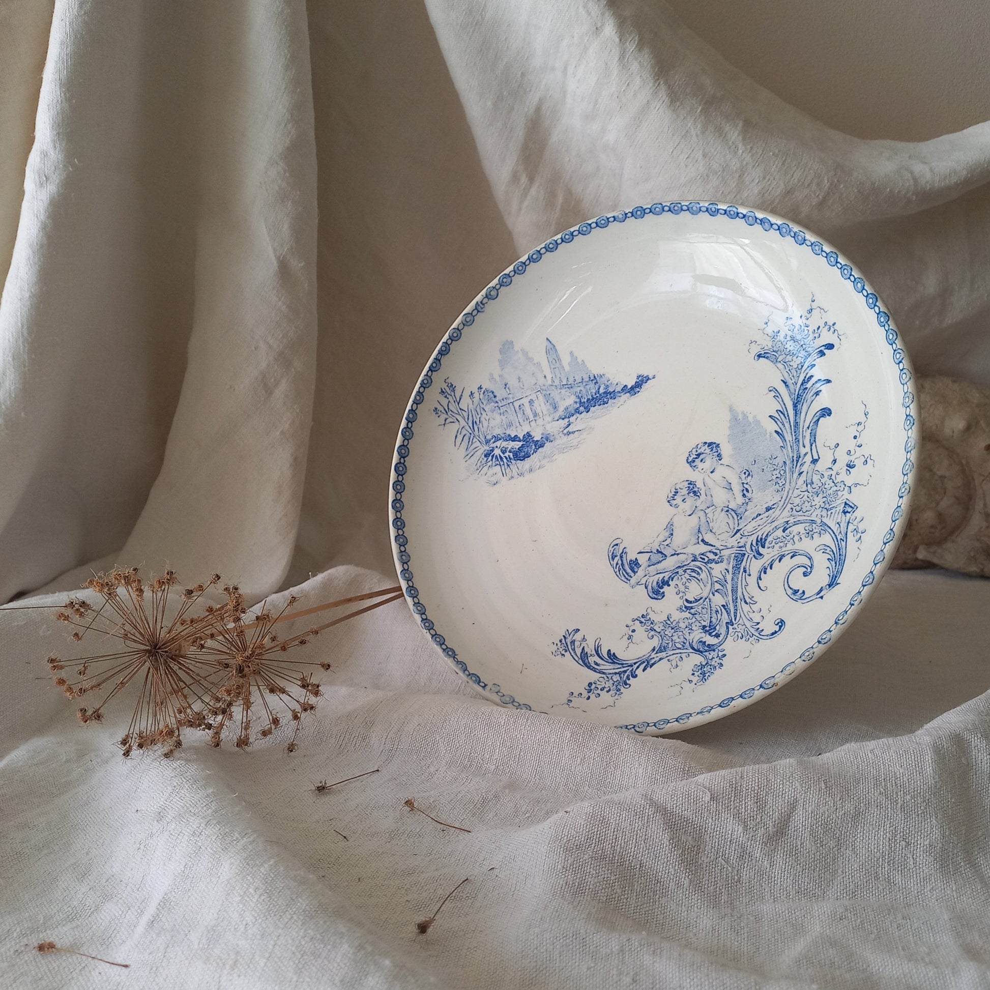 An attractive French antique footed ironstone cake plate with a blue transfer pattern depicting architecture, cherubs, and flowers; a very pretty and delicate pattern.

Original antique ironstone pieces of this age and condition are becoming increasingly difficult to source to this a rare treat to discover.
