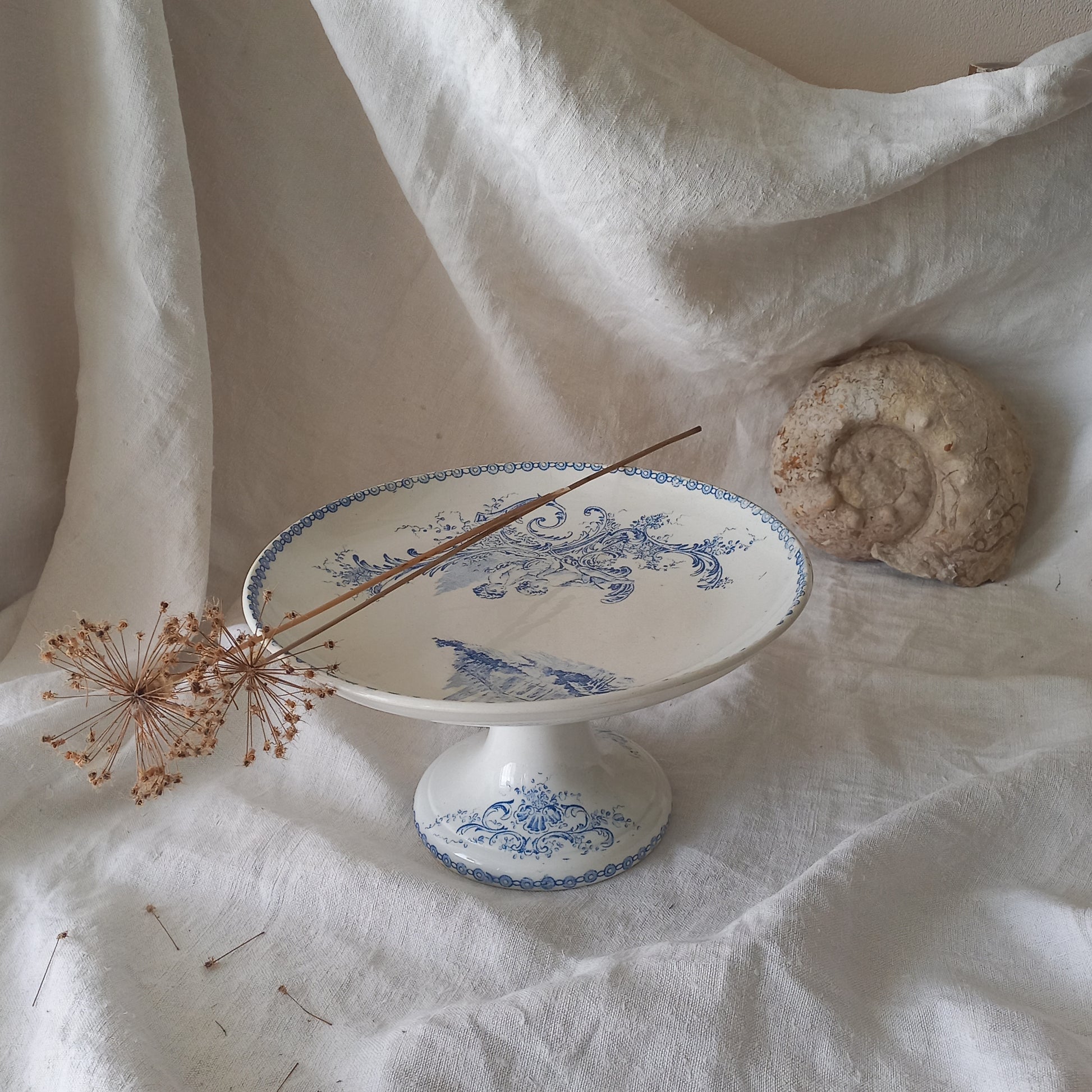 An attractive French antique footed ironstone cake plate with a blue transfer pattern depicting architecture, cherubs, and flowers; a very pretty and delicate pattern.

Original antique ironstone pieces of this age and condition are becoming increasingly difficult to source to this a rare treat to discover.
