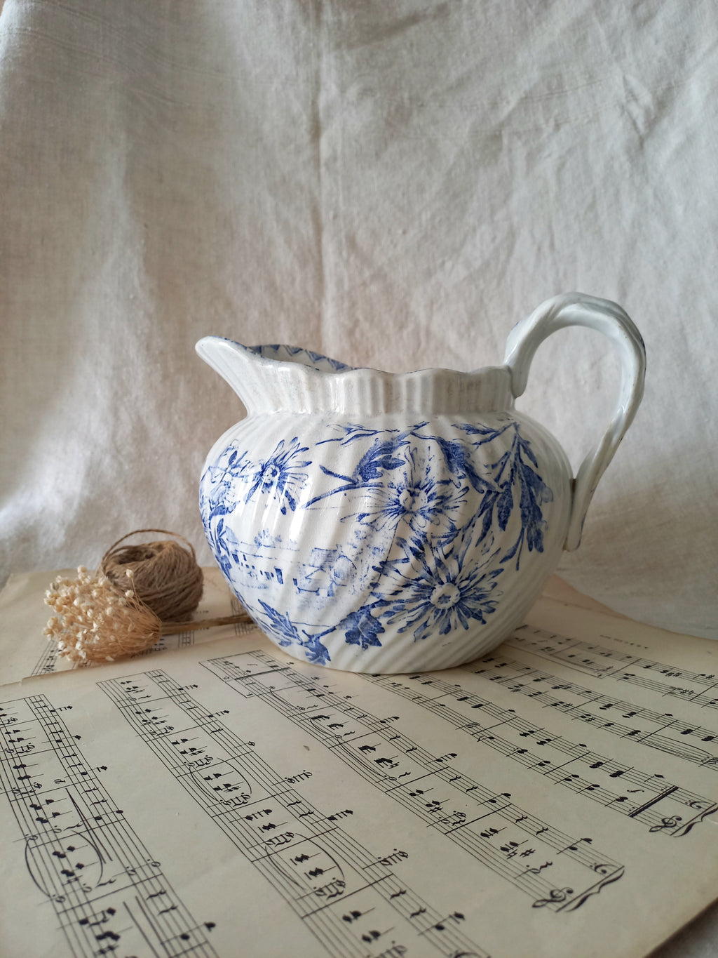 White ceramic pitcher with blue floral patterns on a sheet of music, against a textured white fabric background.