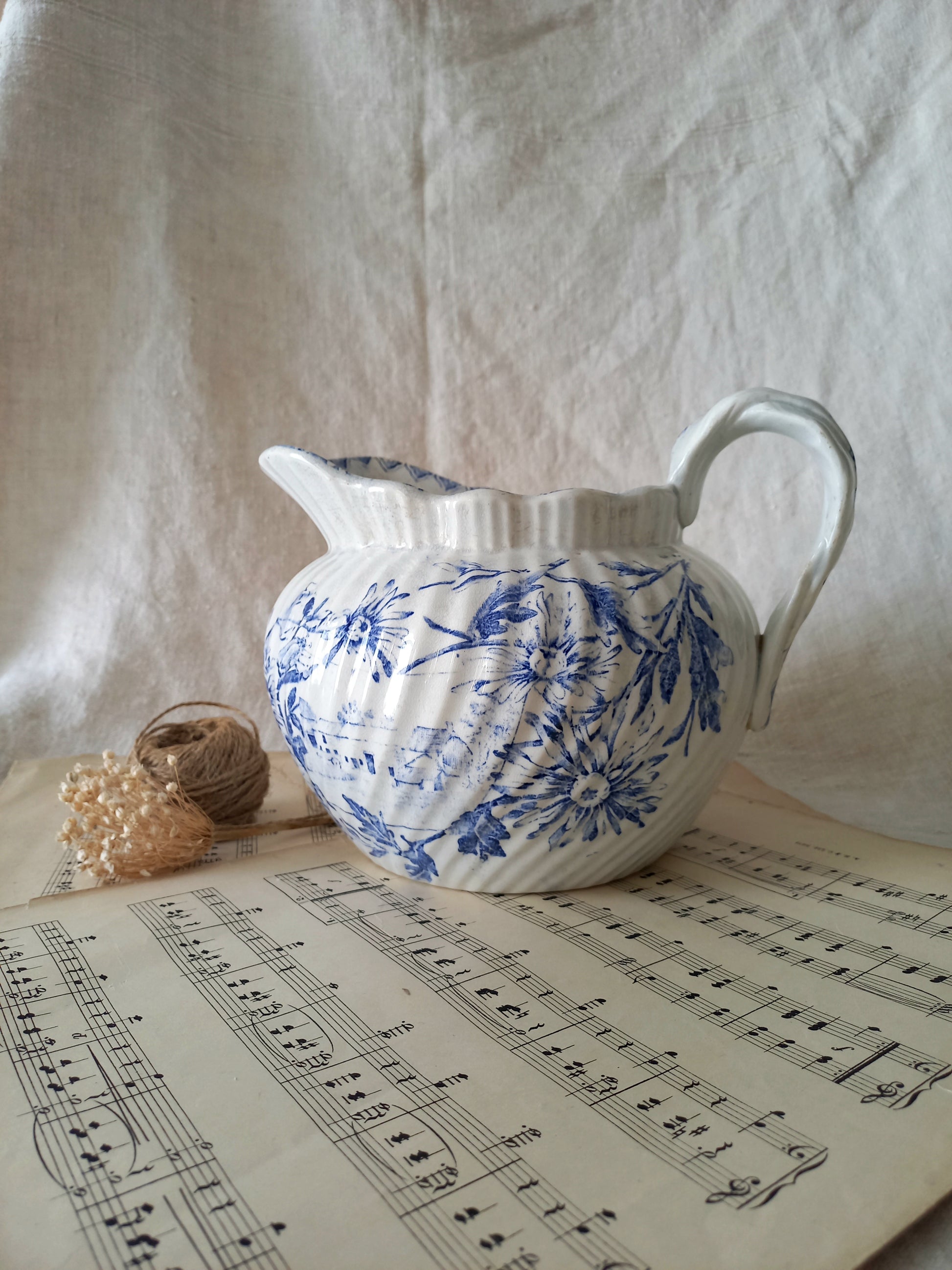 White ceramic pitcher with blue floral patterns on a sheet of music, against a textured white fabric background.