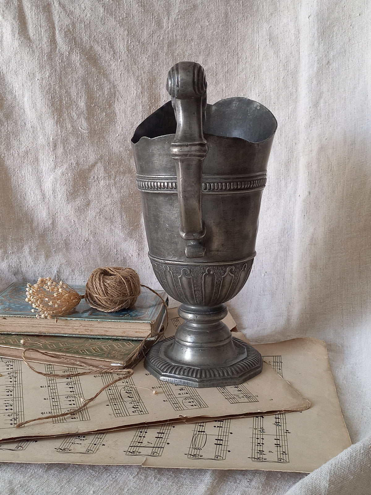 An antique pewter pitcher with a flared base, styled on a textured off-white fabric alongside vintage sheet music, a small shell, and twine.