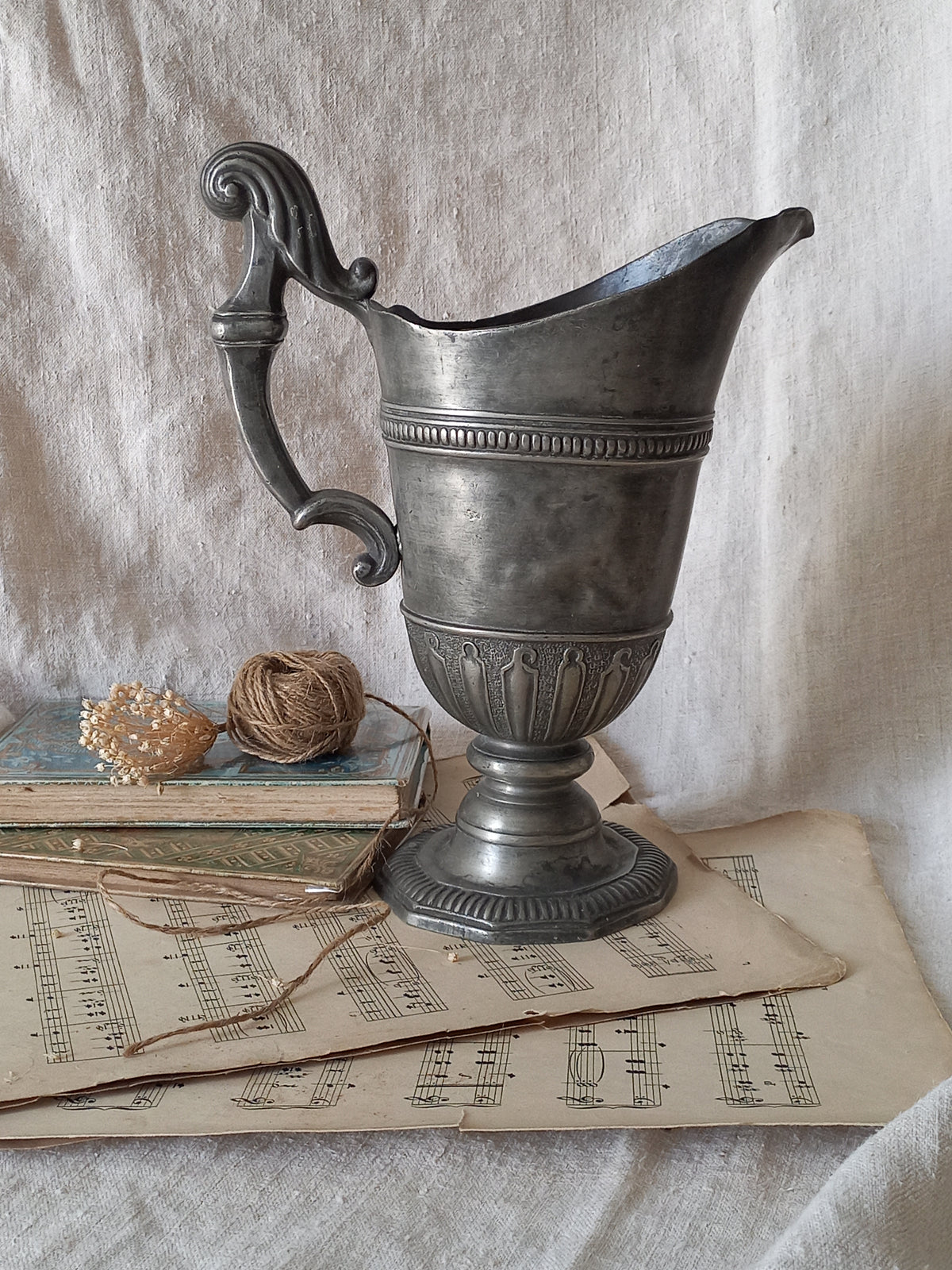 An antique pewter pitcher with a decorative handle, styled on a textured white fabric alongside old sheet music, twine, and a small decorative bead garland.