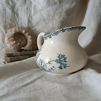 White ceramic pitcher with teal floral patterns on a textured fabric background