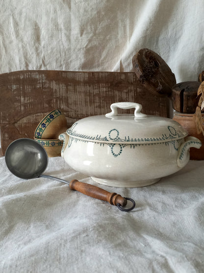 White ceramic tureen with green patterns and a wooden ladle on a white fabric background
