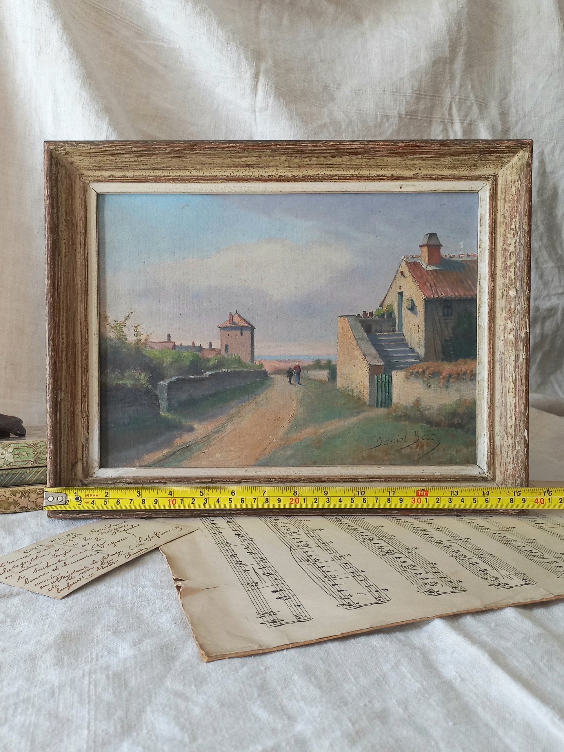 Framed painting of a landscape with houses and a road, placed on a textured surface with music sheets underneath.