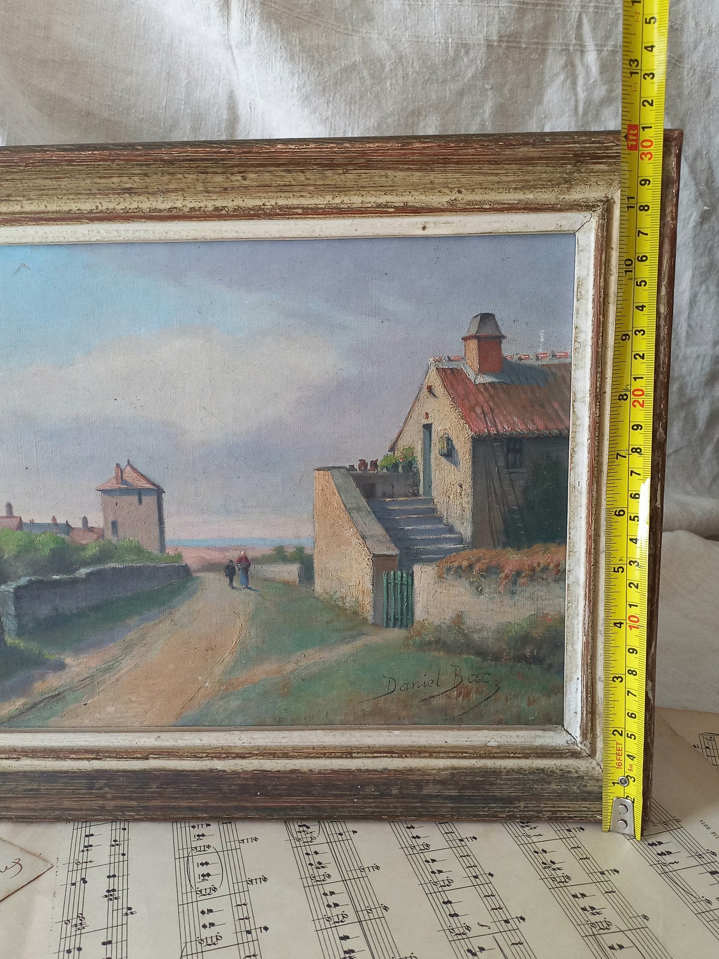 Small oil painting of a village scene with a wooden frame, measured against a white fabric background.
