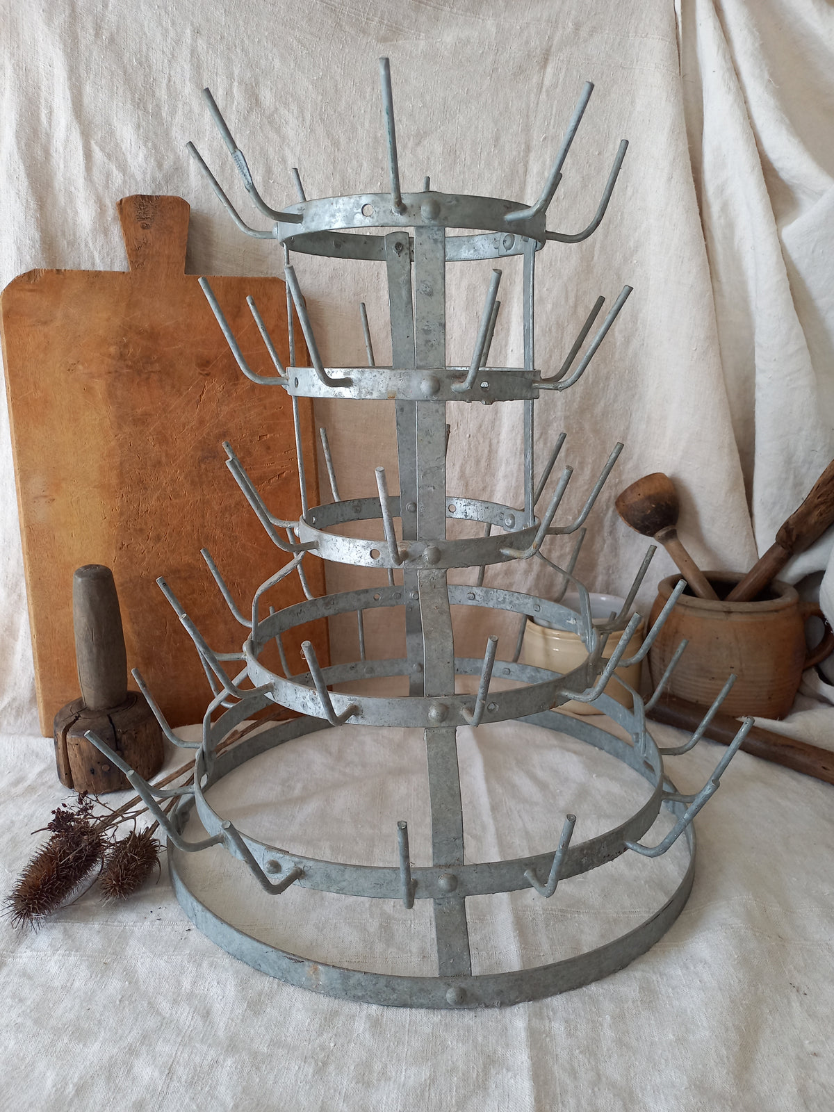 A galvanized metal tiered rack with multiple circular rings and curved hooks, displayed on a draped white fabric backdrop with rustic kitchen props including a wooden cutting board, rolling pin, and wooden spoon holder.