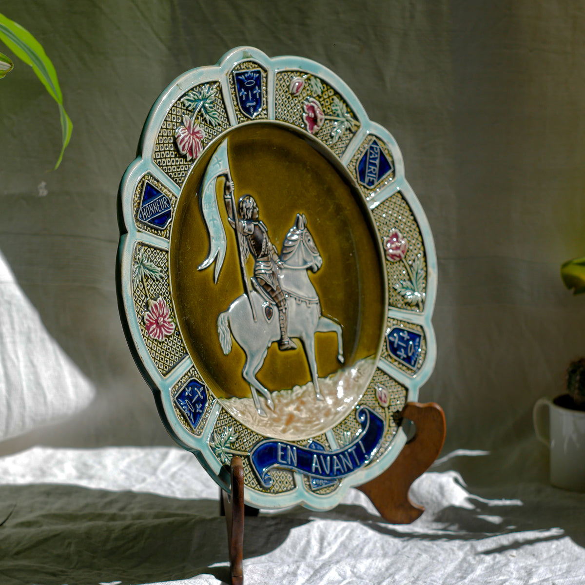 A large vintage/antique majolica wall plate with hand-coloured details in relief, the theme of the plate being the Patron Saint of France - Joan of Arc riding into battle with her banner (in French she is called Jeanne D'Arc).

This wonderful French decorative plate has a rich gloss finish, the glaze even being applied to the underside.

Around the lightly scalloped circumference of this large plate are patriotic symbols such as 'En Avant' (Go Forward), 'Honneur' (Honour) and 'Patrie' (Homeland).
