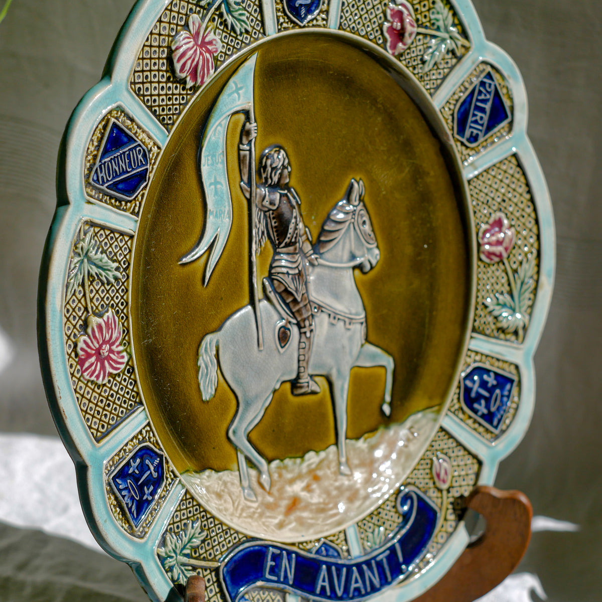 A large vintage/antique majolica wall plate with hand-coloured details in relief, the theme of the plate being the Patron Saint of France - Joan of Arc riding into battle with her banner (in French she is called Jeanne D'Arc).

This wonderful French decorative plate has a rich gloss finish, the glaze even being applied to the underside.

Around the lightly scalloped circumference of this large plate are patriotic symbols such as 'En Avant' (Go Forward), 'Honneur' (Honour) and 'Patrie' (Homeland).