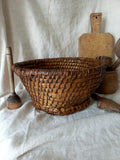 A large round woven vintage basket made of dark brown natural fibers, styled on a draped white linen cloth with rustic wooden kitchen utensils in the background.