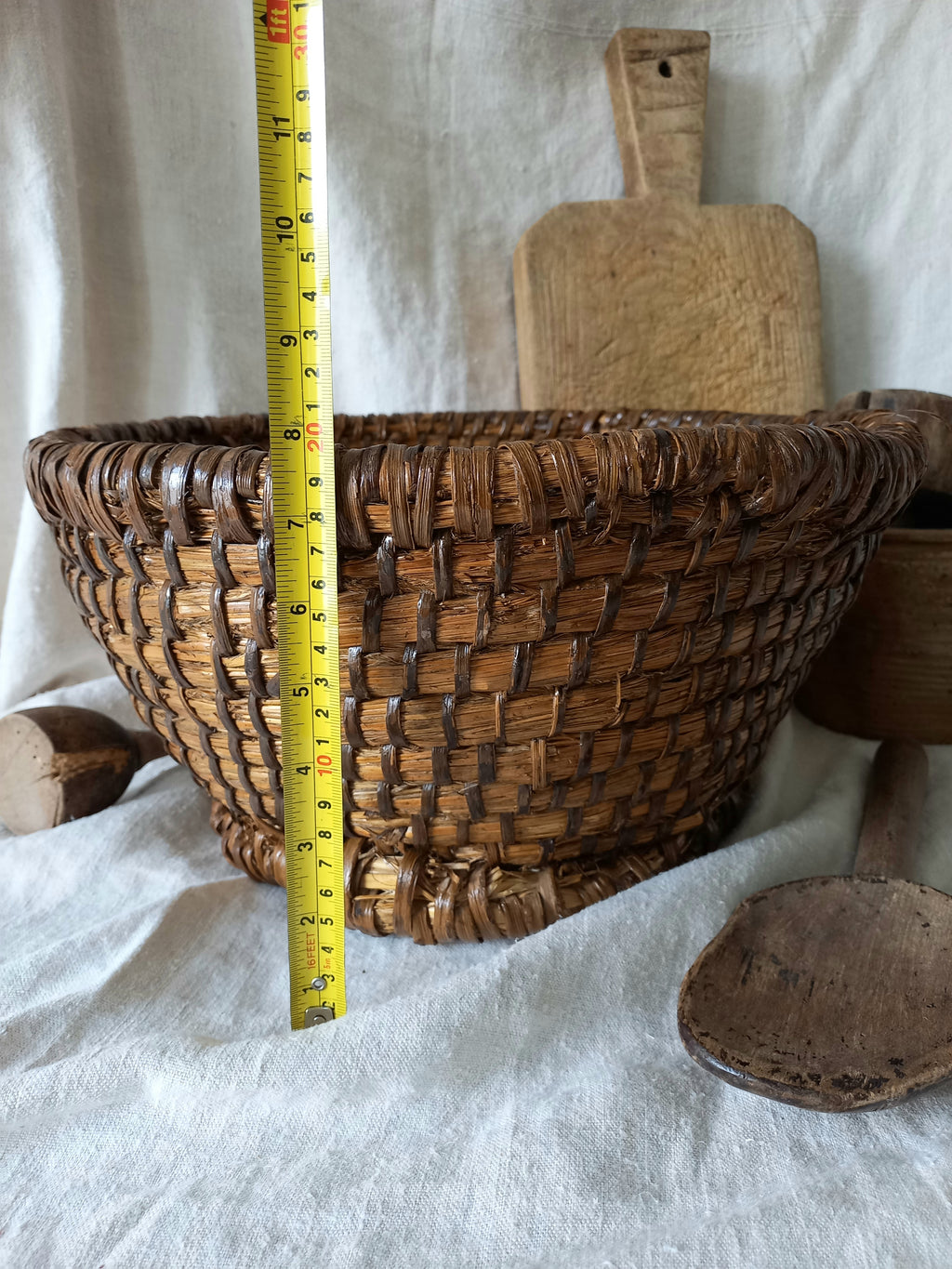 A brown woven vintage basket made from natural fibers, shown with a yellow measuring tape standing vertically next to it for scale, set against a draped white fabric backdrop with rustic wooden props.