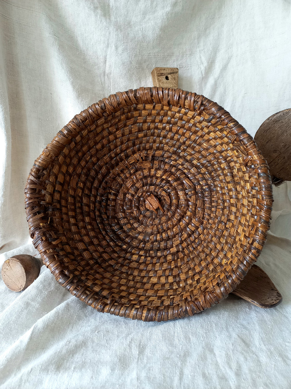 A round woven vintage basket made from natural brown and tan fibers, displayed on a draped white fabric background with rustic wooden props.