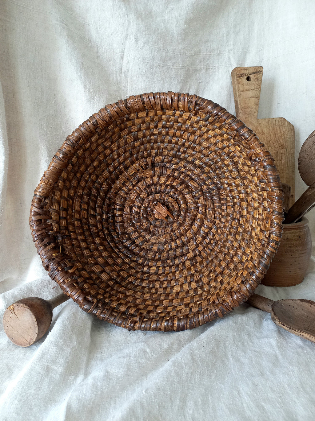 A round, hand-woven brown vintage basket with a spiral pattern, displayed on a draped white linen fabric alongside rustic wooden kitchen utensils.