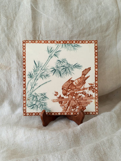A square ceramic tile with a brown sepia illustration of a bird perched on a branch among bamboo leaves, displayed on a small wooden stand against a draped white fabric background.